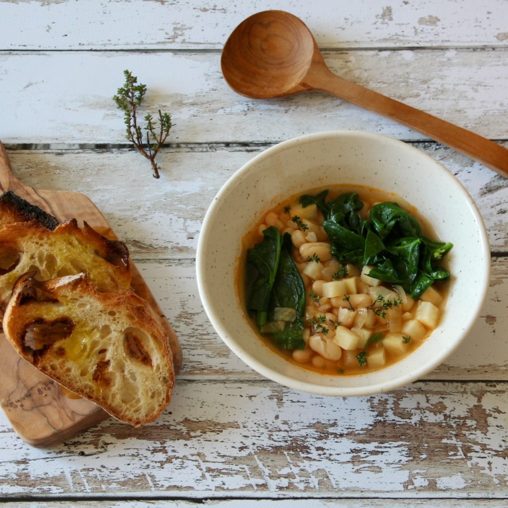 Top view of Tuscan white bean soup with fresh spinach and cubed root vegetables, served with crusty bread on a rustic wooden table