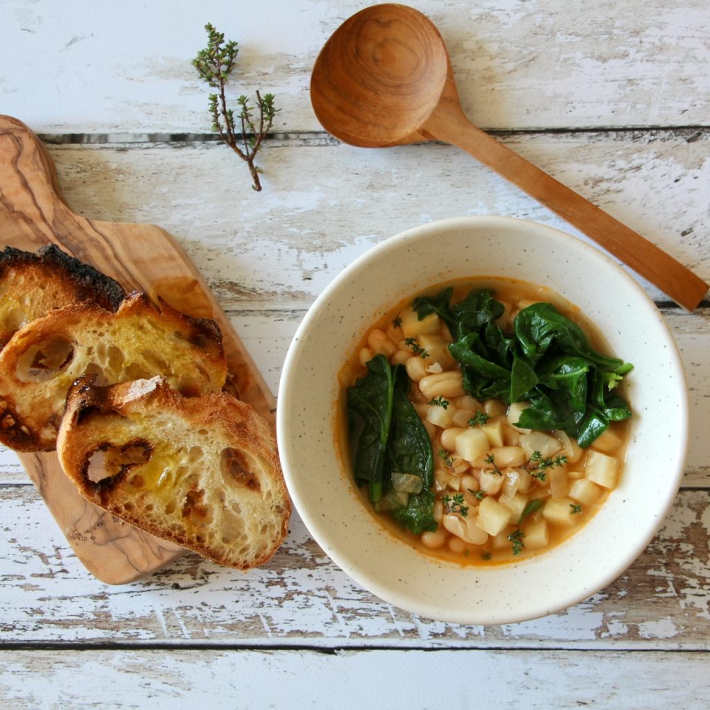 Ingredients for Tuscan white bean soup: fresh spinach, garlic, onions, celeriac, and white beans, displayed on rustic wooden boards